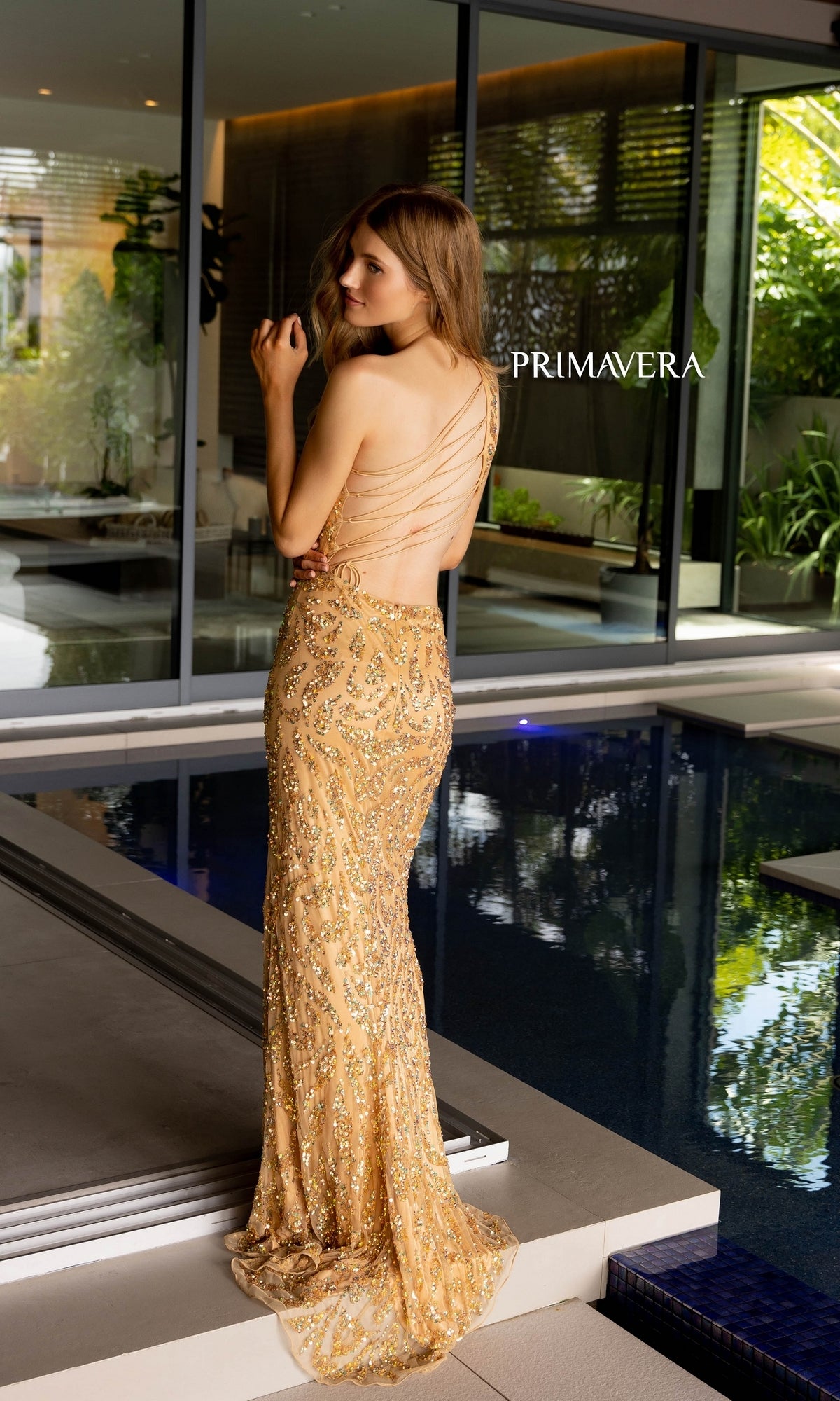 A woman in the Primavera Sequin-Print Long Prom Dress 4191, a gold backless formal gown, stands by an indoor pool facing away.